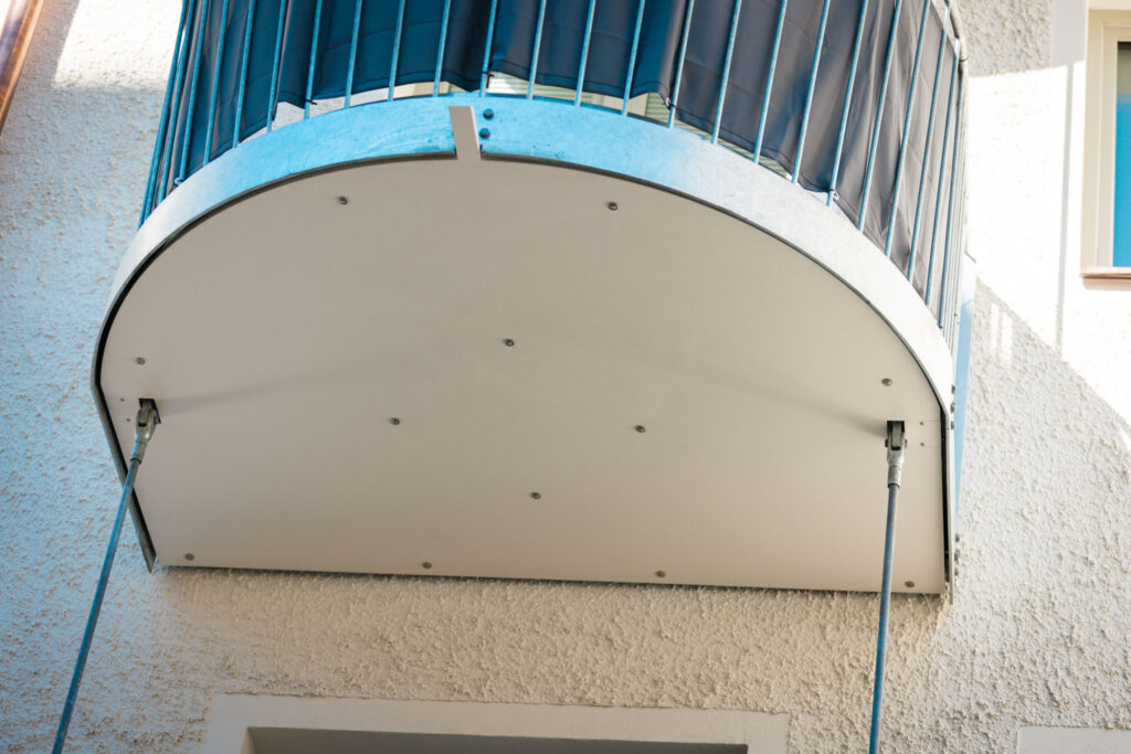 Runder Balkon in Untersicht, mit glatter, weißer Unterverkleidung und sichtbarer Abstützung durch gespannte Drahtseile an der strukturierten Hauswand.