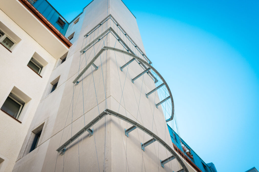 Rundes Metallgeländer an der Außenseite eines hellen Gebäudeturms, als Teil der Struktur für einen Balkon oder eine Rankhilfe, vor leuchtend blauem Himmel.