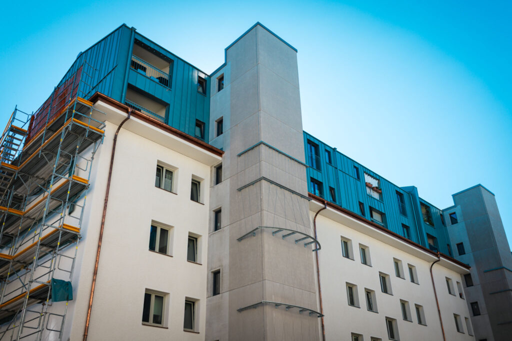Ansicht des Mehrfamilienhauses mit mehreren kleinen, runden Balkonen an der hellen Hauptfassade und einem turmartigen Anbau in der Mitte.