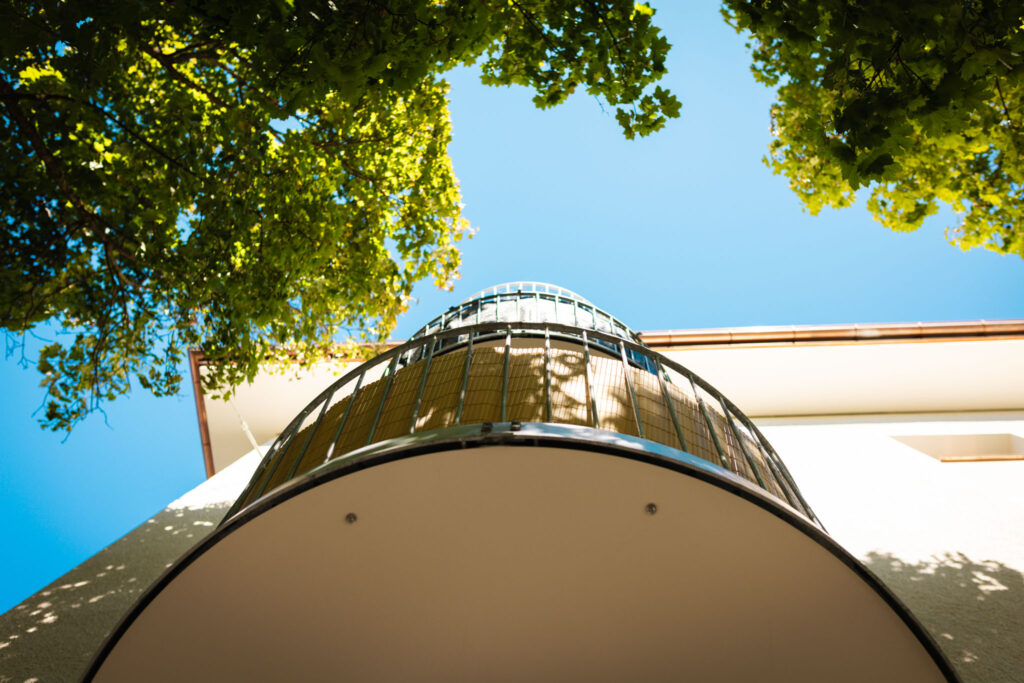 Runder Balkon in extremer Untersicht, wobei das Metallgeländer und die innere Verkleidung gegen den klaren blauen Himmel und das umgebende grüne Laub betont werden.