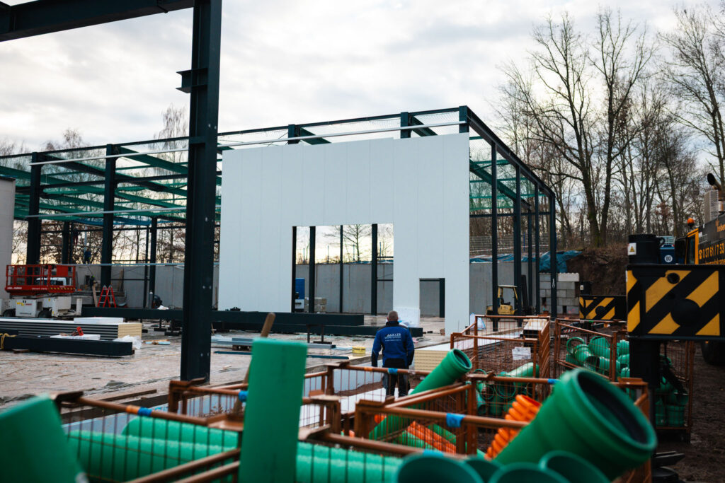 Nahaufnahme eines im Bau befindlichen Hallengebäudes in Stahlrahmenbauweise. Ein großes, weißes Wandelement mit einer Fensteröffnung wird in die Metallkonstruktion eingesetzt. Im Vordergrund sind grüne und orangefarbene Rohre oder Kabelrollen zu sehen.