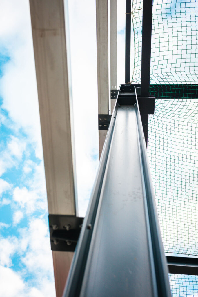 Detailaufnahme, die vertikal nach oben blickt. Sie zeigt einen massiven, schwarzen Stahlträger der Konstruktion vor einem hellblauen Himmel mit weißen Wolken. Rechts ist ein grünes Sicherheitsnetz sichtbar.