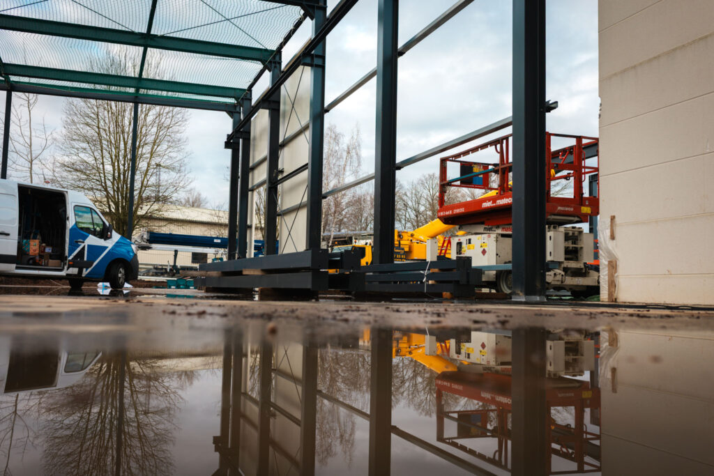Eine Szene auf der Baustelle an einem bewölkten Tag, bei der sich der Stahlrahmen und die umgebenden Bäume deutlich in einer großen Pfütze auf dem Baustellengelände spiegeln. Ein gelber Kran oder eine Hubarbeitsbühne steht im Hintergrund.