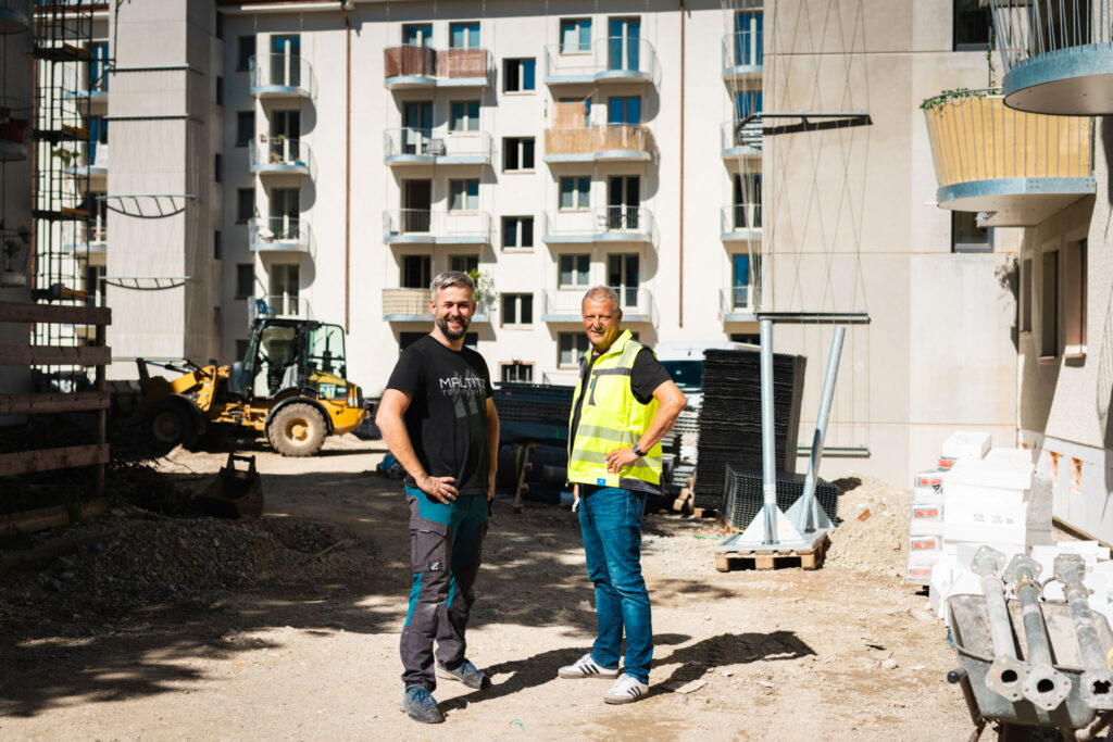 Porträt von Frank Maltitz und Alexander Fritzsch, zueinandergedreht und mit den Hnden in den Hüften, einer in Warnweste, vor der hellen Fassade mit neuen, runden Balkonen im Hintergrund.