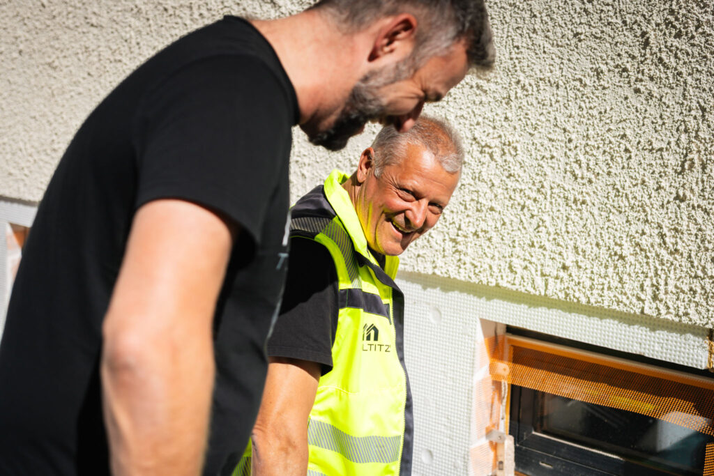 Zwei lächelnde Mitarbeiter von Maltitz, Alexander Fritzsch und Frank Maltitz, oder Bauleiter in Arbeitskleidung, einer davon in leuchtender Warnweste, besprechen Details an einer unfertigen Fassade mit Dämmmaterial.
