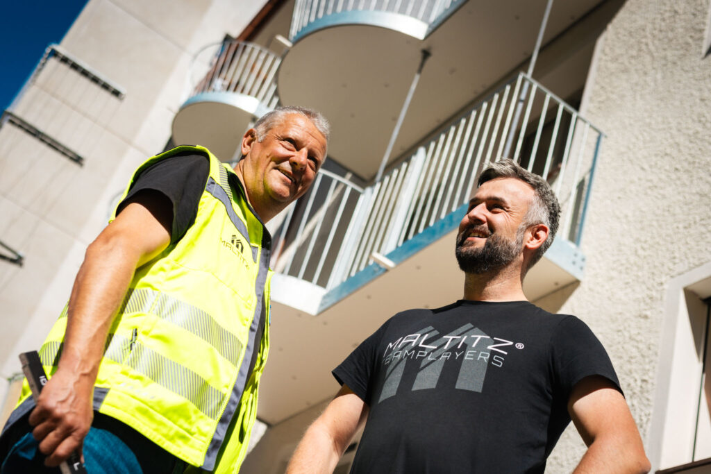 Porträt von zwei lachenden Bauleitern oder Handwerkern, einer in Warnweste, vor der hellen Fassade mit neuen, runden Balkonen im Hintergrund.