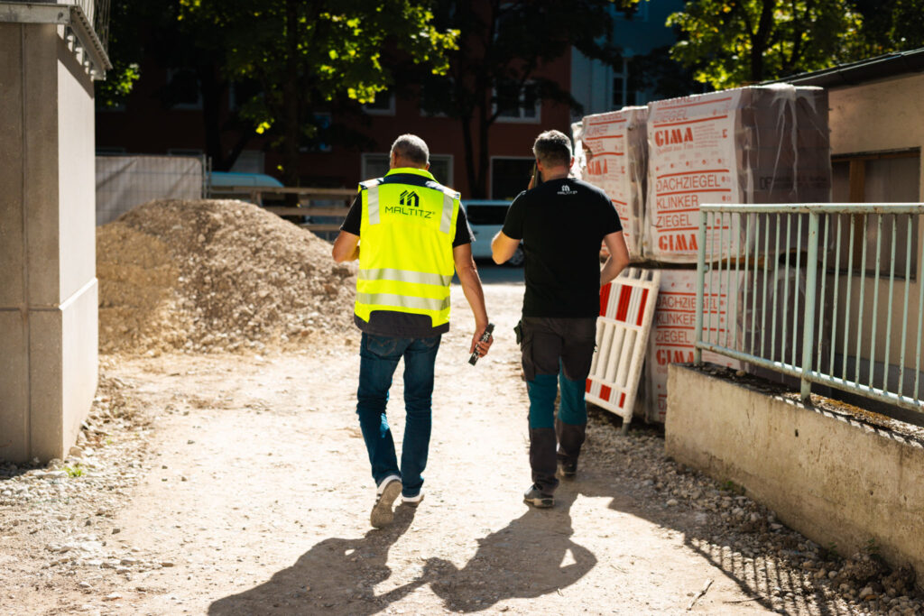 Zwei Bauarbeiter oder Bauleiter, einer in gelber Warnweste, gehen von der Kamera weg auf einem Baugelände entlang, vorbei an einer Schotterhalde und Baumaterialien.