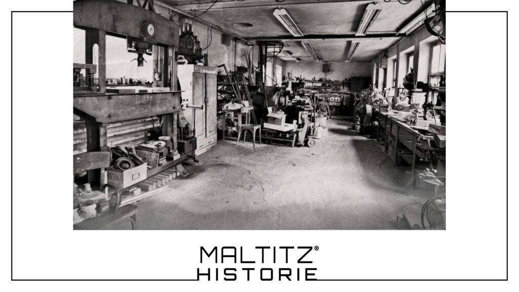 Schwarz-Weiß-Aufnahme der historischen Werkstatt von Maltitz um 1913–1940. Zu sehen ist eine traditionelle Bauschlosserei mit einer großen mechanischen Presse, Werkzeugschränken und Werkbänken in einer authentischen Industriehalle.