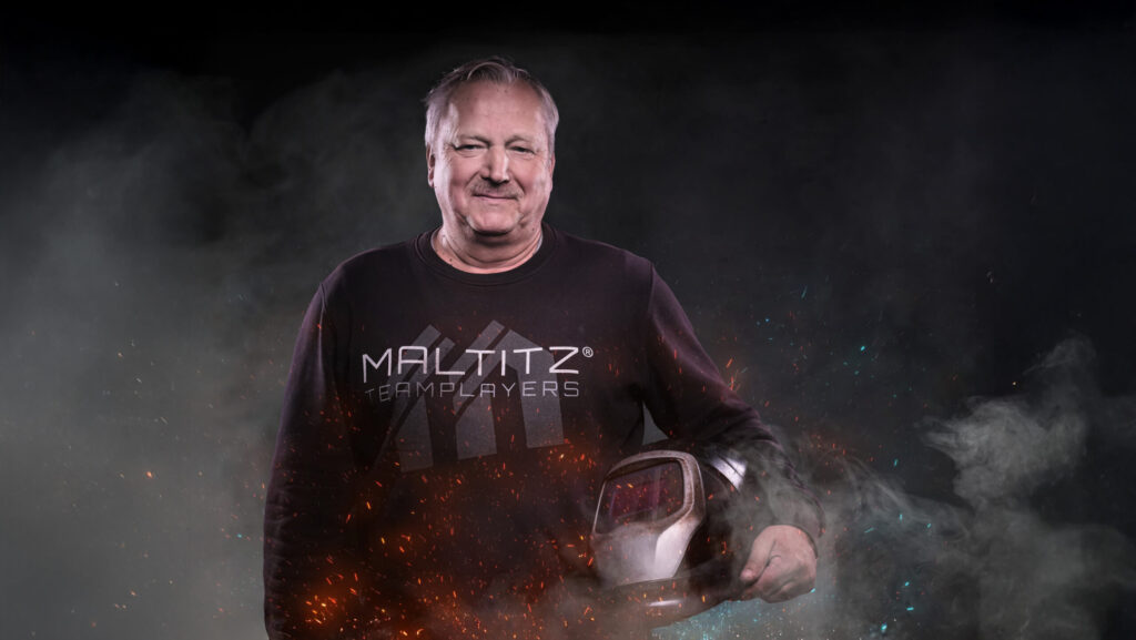 Lächelnder Mitarbeiter Michael von MALTITZ Metallbau mit Schweißerhelm unter dem Arm. Funken sprühen, Rauch steigt auf.