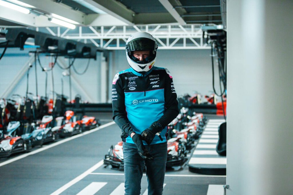 Portrait von Dirk Maltitz in Rennfahrerausstattung, mit Helm und türkisem CFMoto-Oberteil. Er zieht sich gerade einen Handschuh an, während er im Vordergrund einer Reihe Karts posiert.