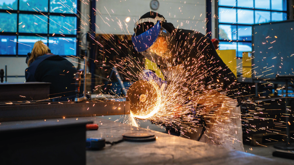 Nahaufnahme einer Person in Schutzkleidung, die mit einem Winkelschleifer Metall bearbeitet, wobei ein intensiver Funkenregen in einem dunklen Werkstattbereich entsteht.