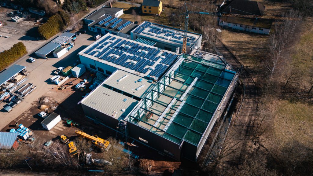 Drohnenaufnahme, die das gesamte Gelände plus neu gebaute hallen aus der Luft zeigt Bei der kleineren Halle ist das fast fertige Dach zu sehen, die größere Halle hat bereits die Unterkonstruktion drauf.