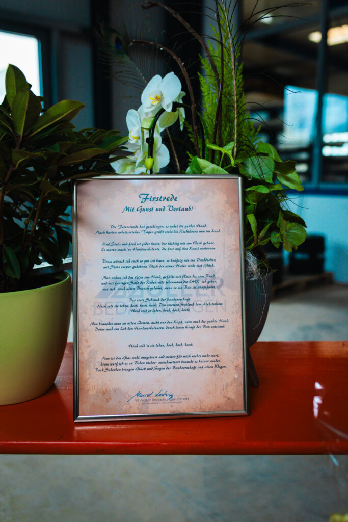 Gerahmte „Firstrede“ (Richtspruch) auf einem Tisch neben Blumenarrangements als Teil der feierlichen Tradition.