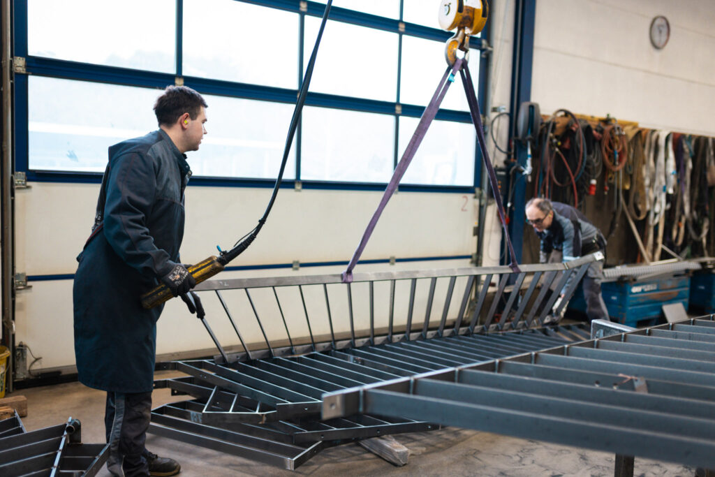 Zwei Metallbauer bewegen ein langes Geländersegment mithilfe eines Krans sicher durch die Werkstatt.