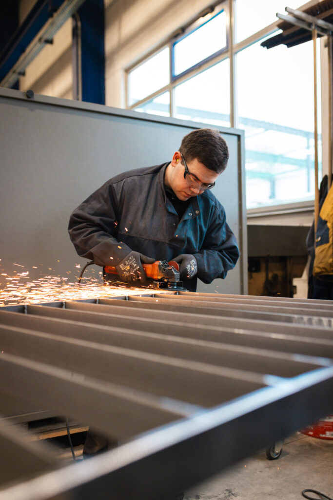 Ein Metallbauer bearbeitet die Schweißnähte eines Geländers mit einem Winkelschleifer, während Funken sprühen.