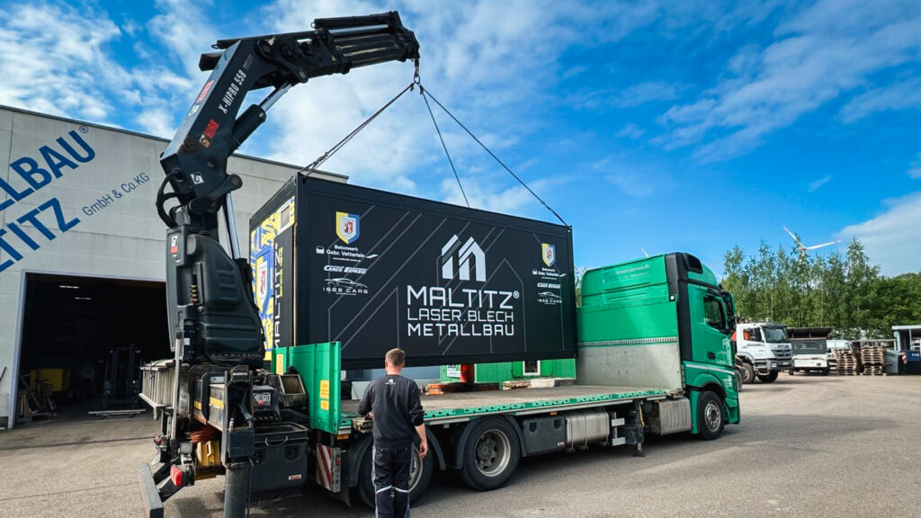 Ein großer grüner Lkw der Spedition Pross mit einem Ladekran lädt auf dem Gelände von Maltitz Metallbau eine schwarze Container-Foodbox auf die Ladefläche. Auf der Box ist groß das Logo von Maltitz Laser Blech Metallbau sowie das Vereinswappen des SV Waldenburg zu sehen. Ein Mitarbeiter überwacht den Vorgang.