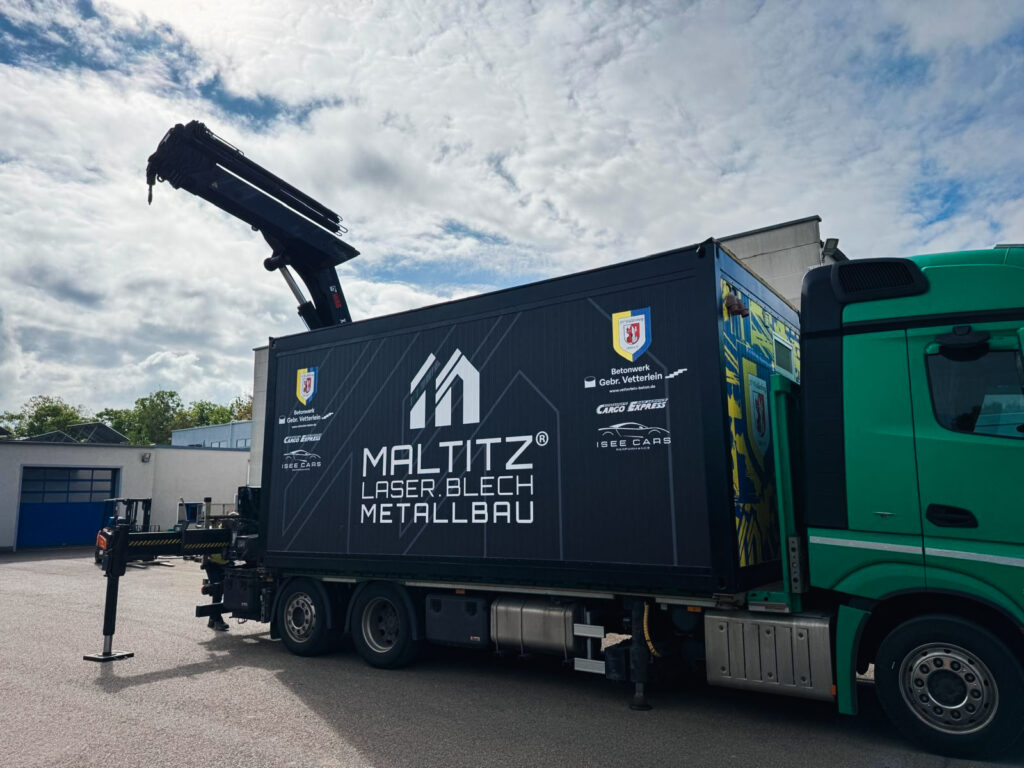 Seitenansicht des grünen Lkw mit der fertig aufgeladenen, schwarzen Foodbox (Schmecktempel). Die Seitenwand der Box zeigt das weiße Maltitz-Logo und Sponsoren-Logos wie das des SV Waldenburg. Der Kranarm des Lastwagens ist über der Box eingeklappt.