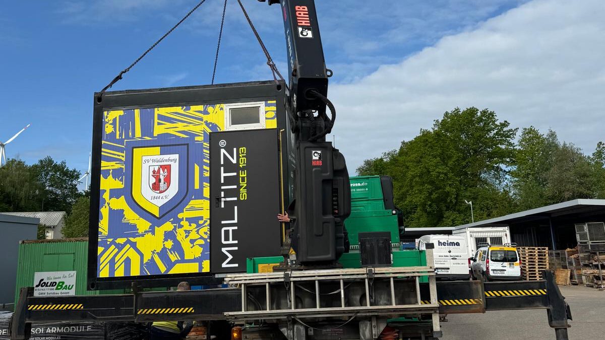 Rückansicht des Lkw mit ausgefahrenen Stützen. Der Kran hebt die Verpflegungsbox an. Die auffällige gelb-blaue Stirnseite der Box trägt das Vereinswappen des SV Waldenburg 1844 e.V. und den Schriftzug „MALTITZ SINCE 1913“.