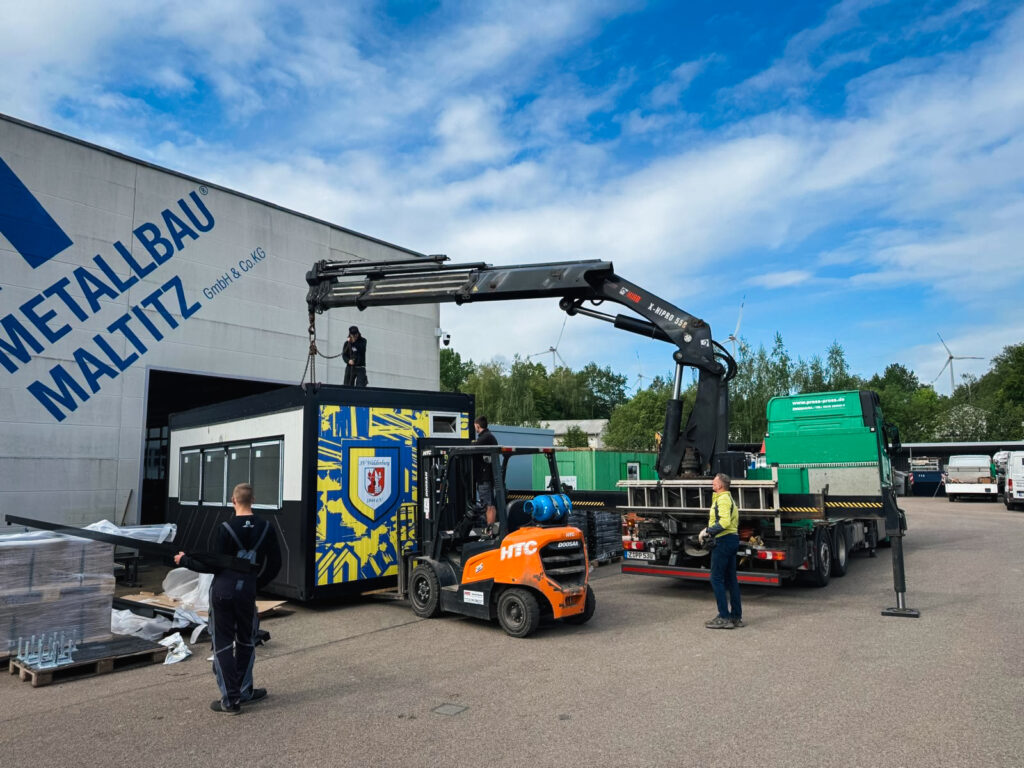 Verladearbeiten auf dem Betriebsgelände: Ein orangefarbener Gabelstapler und der Kran des Lkw positionieren gemeinsam die moderne Foodbox. Sichtbar ist hier die farbenfrohe gelb-blaue Rückseite des Containers mit dem großen Wappen des SV Waldenburg. Ein Arbeiter steht auf dem Dach der Box, um die Ketten zu sichern.
