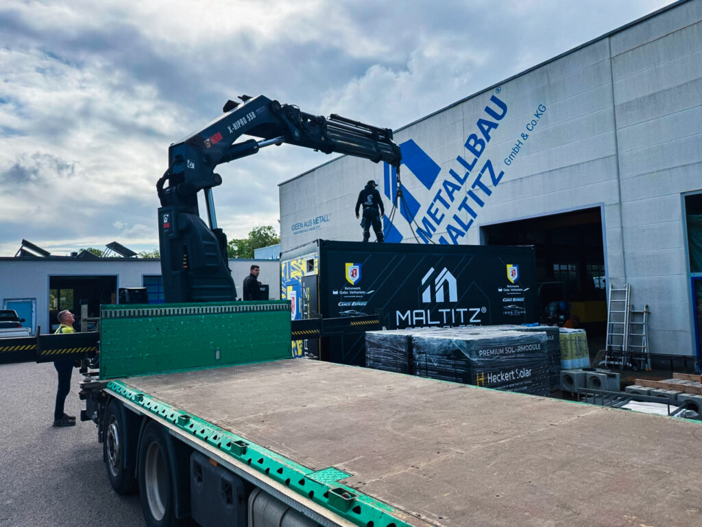 Blick von der leeren Ladefläche des Lkw in Richtung der Werkshalle von Metallbau Maltitz. Im Hintergrund steht die schwarze Foodbox mit Sponsorenaufdrucken, während ein Mitarbeiter auf dem Dach des Containers die Hebeketten des Ladekrans befestigt.