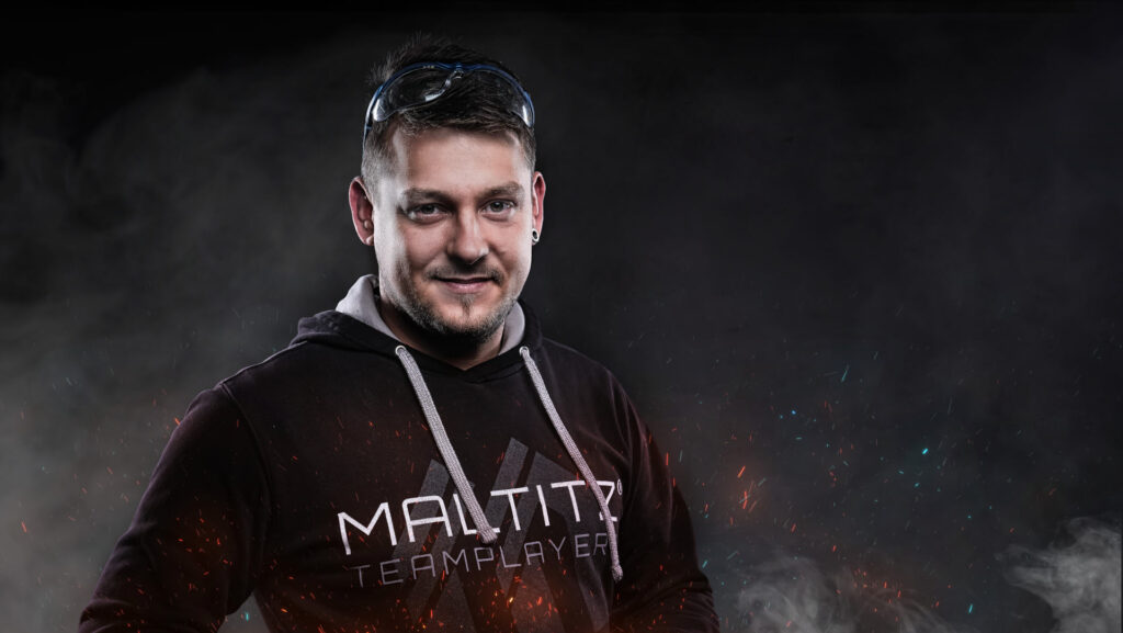 MALTITZ Teamplayers - Vorstellung Patrick Ein junger Mann mit Maltitz-Pullover steht vor einem grauen Hintergrund, um ihn herum Funken