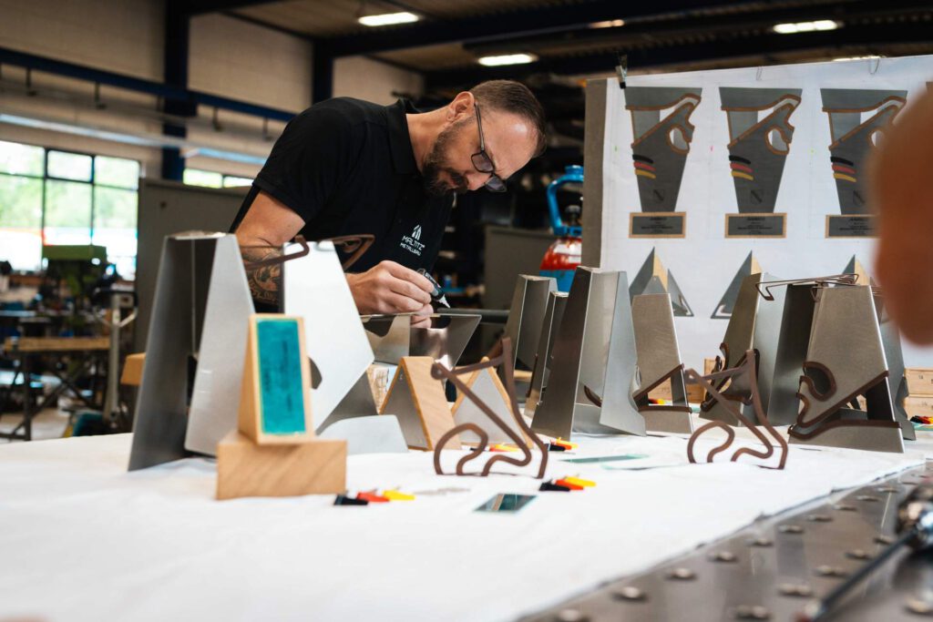 Ein Handwerker in der Werkhalle bei der präzisen Montage und Veredelung mehrerer MotoGP-Trophäen auf einem Arbeitstisch mit Konstruktionszeichnungen im Hintergrund.