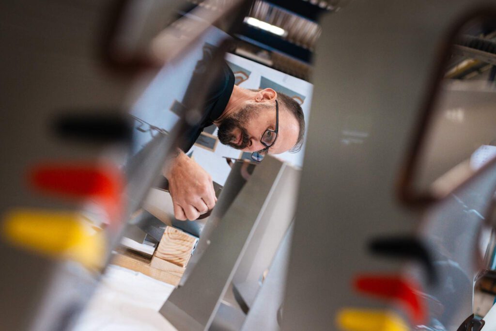 Blick durch die Pokal-Elemente auf einen Metallbauer mit Brille, der konzentriert per Hand die Details der Trophäen für den Motorrad Grand Prix anpasst.