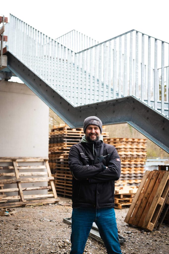 Ein lächelnder Mitarbeiter von Metallbau Maltitz steht mit verschränkten Armen vor einer montierten Fluchttreppen-Konstruktion.
