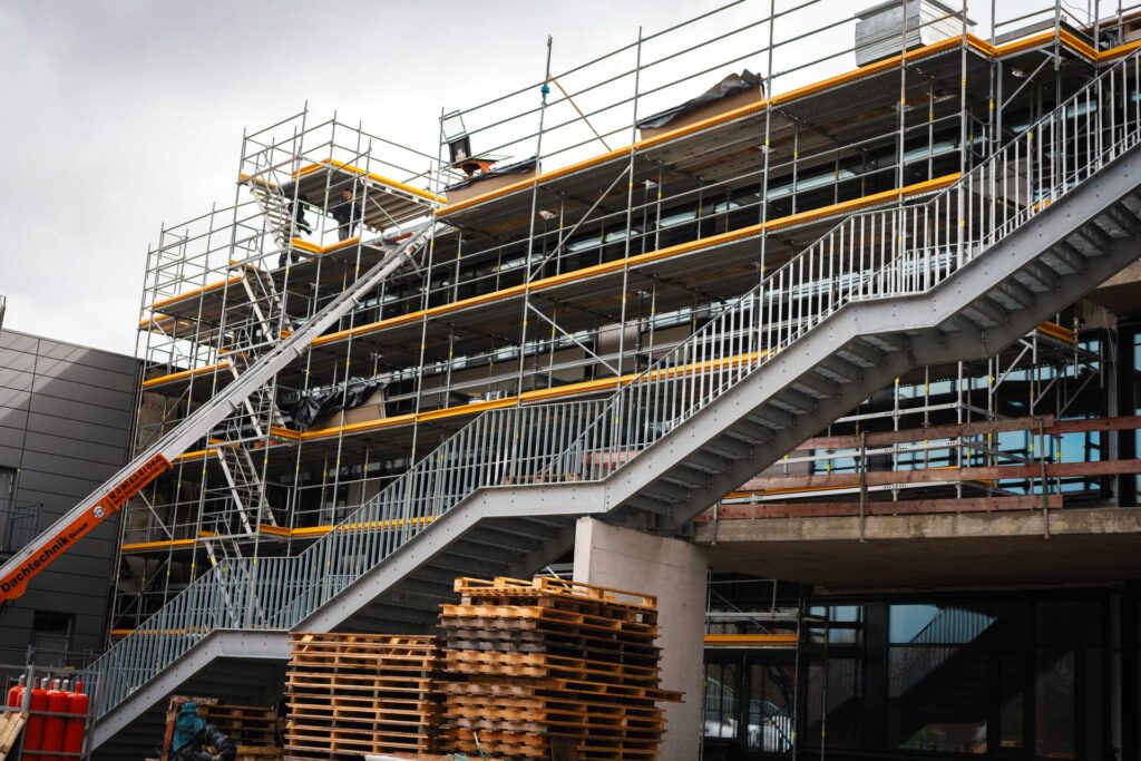 Großansicht der Baustelle in Haimhausen mit Kranarbeiten für die Montage großer Fluchttreppen und Fluchtbalkone.