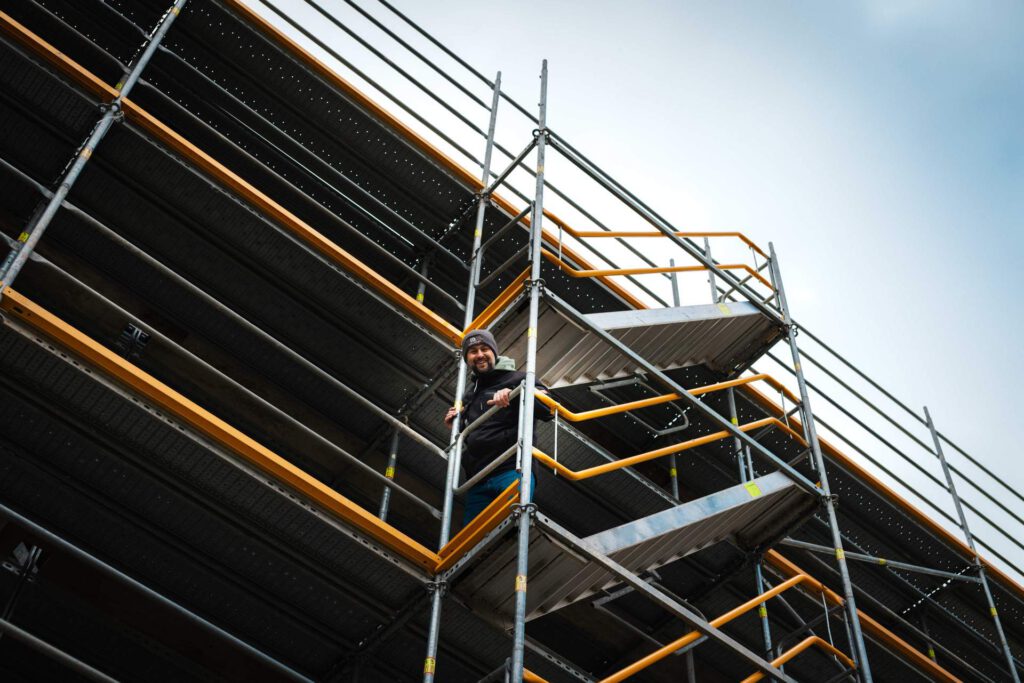 Ein lächelnder Mitarbeiter von Metallbau Maltitz steht gesichert auf einem hohen Baugerüst an der Fassade in Haimhausen.
